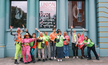 A group of people in colorful outfits wave and pose in front of a building with "UNION STREET PARTY" written on the window. Photo taken by Hanna Collins Photography for the Union Street Party organised by Nudge Community Builders