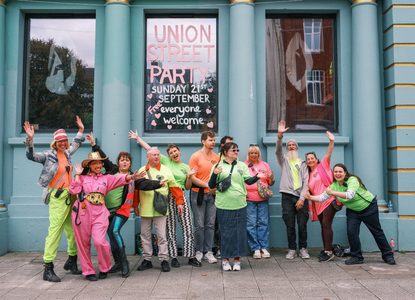 A group of people in colorful outfits wave and pose in front of a building with "UNION STREET PARTY" written on the window. Photo taken by Hanna Collins Photography for the Union Street Party organised by Nudge Community Builders