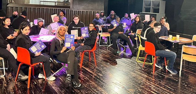 Young people sat around small tables holding up a training booklet at a Good Night Out Campaign training session
