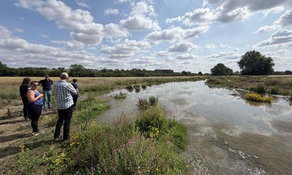 Text over image saying: New blog - Land truths, when the fires are burning. Image below it shows a small group of people stand beside a shallow river, surrounded by low green shrubs and grass. Picture taking on an Esmée Fairbairn Foundation visit to the E