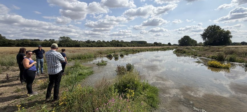 Text over image saying: New blog - Land truths, when the fires are burning. Image below it shows a small group of people stand beside a shallow river, surrounded by low green shrubs and grass. Picture taking on an Esmée Fairbairn Foundation visit to the E