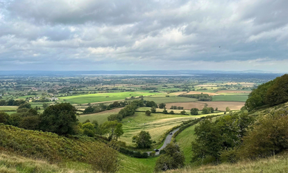 View from a hillside over a patchwork countryside of green fields and hedgerows, with a winding road through trees and distant hills beneath cloudy skies-Stroud
