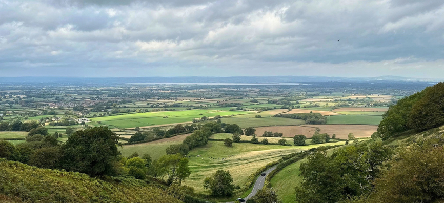 View from a hillside over a patchwork countryside of green fields and hedgerows, with a winding road through trees and distant hills beneath cloudy skies-Stroud